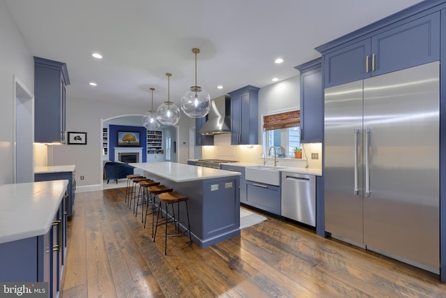 kitchen with a sink, blue cabinetry, stainless steel appliances, wall chimney exhaust hood, and a fireplace