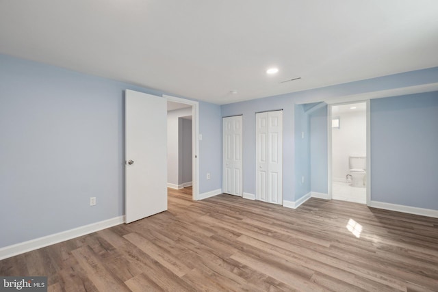 unfurnished bedroom with two closets, ensuite bathroom, wood finished floors, recessed lighting, and baseboards