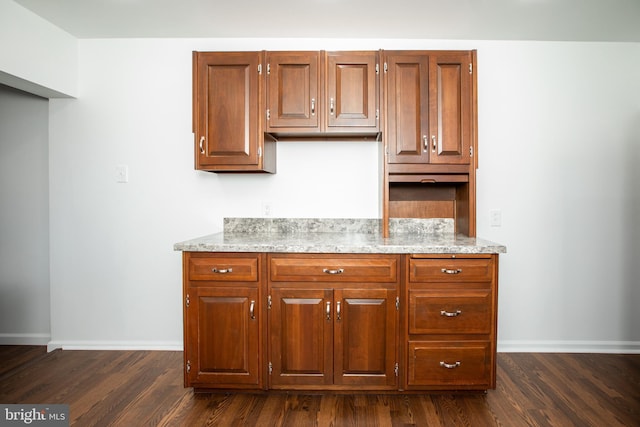 kitchen with brown cabinetry, dark wood finished floors, light stone countertops, and baseboards