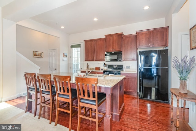 kitchen with a kitchen bar, black appliances, an island with sink, wood finished floors, and recessed lighting