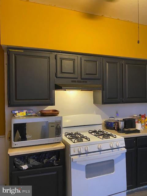 kitchen with under cabinet range hood, white range with gas cooktop, light countertops, dark cabinetry, and stainless steel microwave