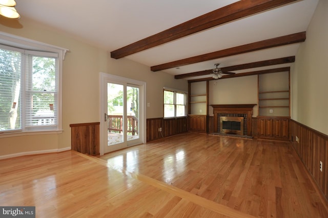 unfurnished living room with a ceiling fan, light wood-type flooring, wainscoting, beam ceiling, and a brick fireplace