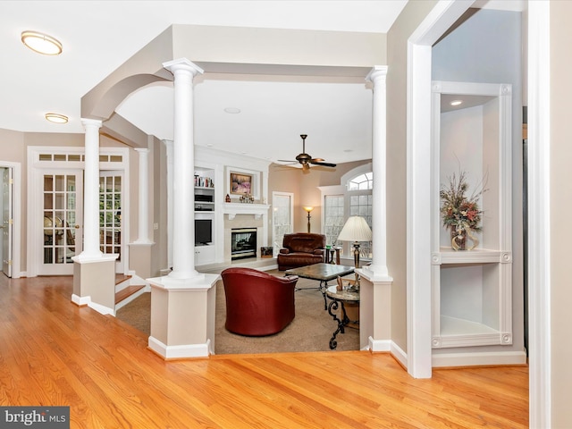 living room with wood finished floors, a glass covered fireplace, a ceiling fan, and ornate columns