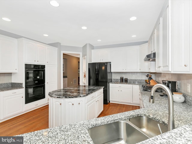 kitchen featuring under cabinet range hood, a sink, light wood-type flooring, dark stone counters, and black appliances