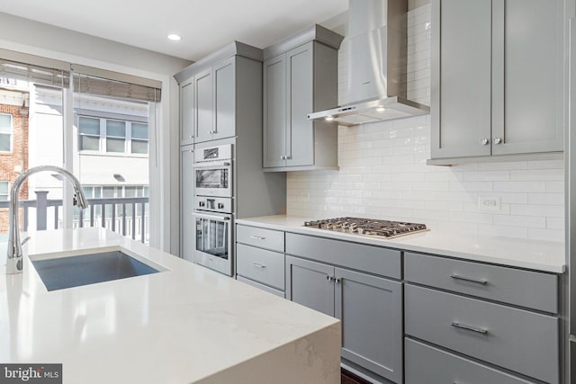 kitchen with gray cabinets, a sink, decorative backsplash, appliances with stainless steel finishes, and wall chimney exhaust hood