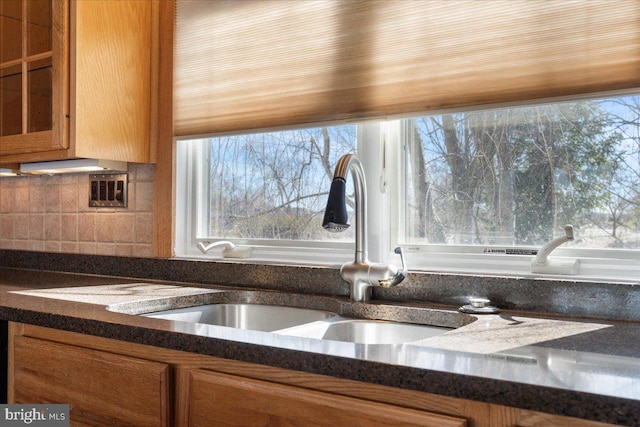 kitchen with a sink, brown cabinets, decorative backsplash, dark stone countertops, and glass insert cabinets