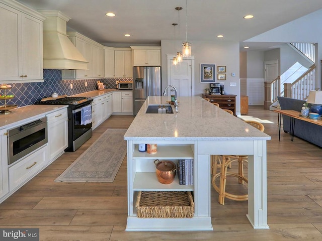 kitchen with open shelves, stainless steel appliances, a kitchen island with sink, a sink, and premium range hood