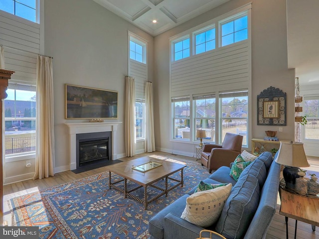 living room featuring coffered ceiling, wood finished floors, a fireplace with flush hearth, a towering ceiling, and a healthy amount of sunlight