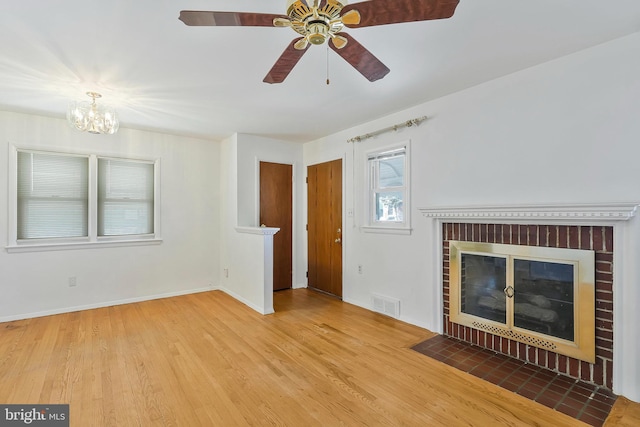 unfurnished living room with baseboards, visible vents, wood finished floors, a brick fireplace, and ceiling fan with notable chandelier