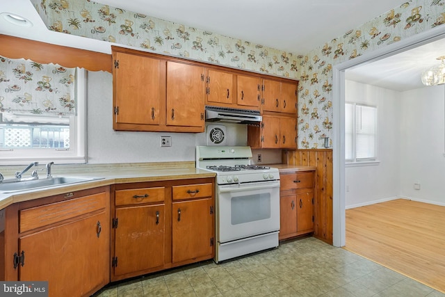 kitchen with brown cabinets, a sink, white gas range oven, under cabinet range hood, and wallpapered walls