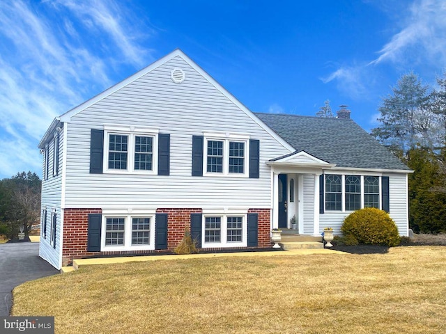 split level home with aphalt driveway, brick siding, roof with shingles, a chimney, and a front lawn