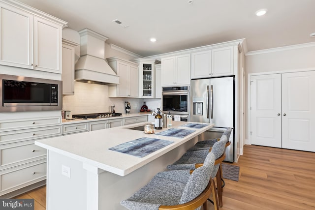 kitchen featuring light wood finished floors, custom range hood, appliances with stainless steel finishes, a kitchen bar, and crown molding