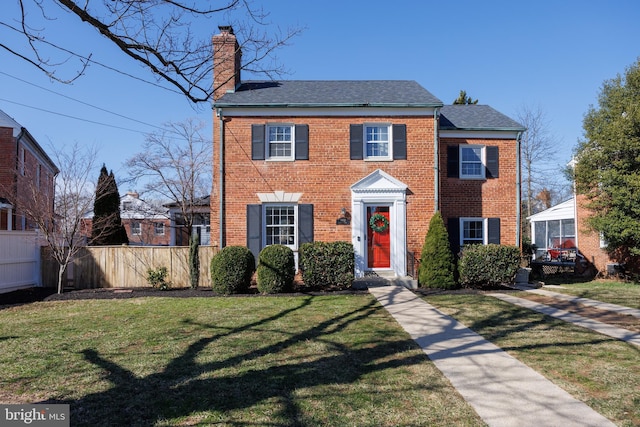 colonial-style house featuring brick siding, a chimney, a front yard, and fence