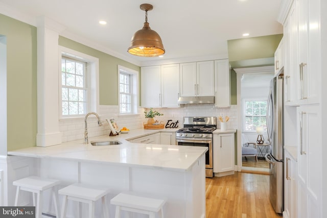 kitchen with a peninsula, a sink, under cabinet range hood, appliances with stainless steel finishes, and white cabinetry