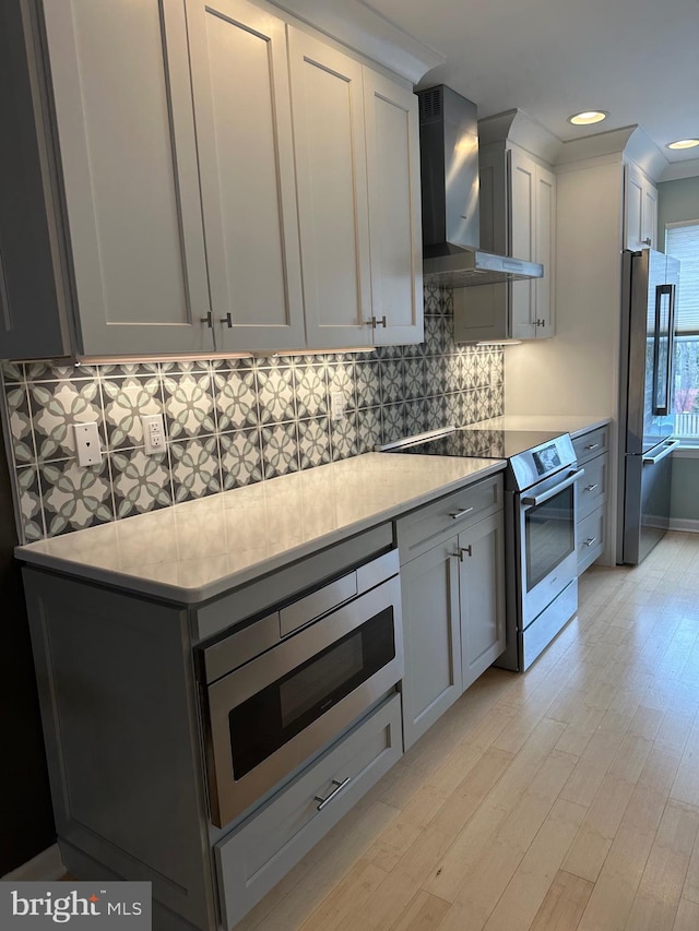 kitchen with light wood finished floors, backsplash, wall chimney range hood, recessed lighting, and appliances with stainless steel finishes