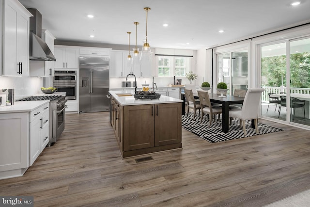 kitchen with high end appliances, an island with sink, wall chimney exhaust hood, a sink, and backsplash