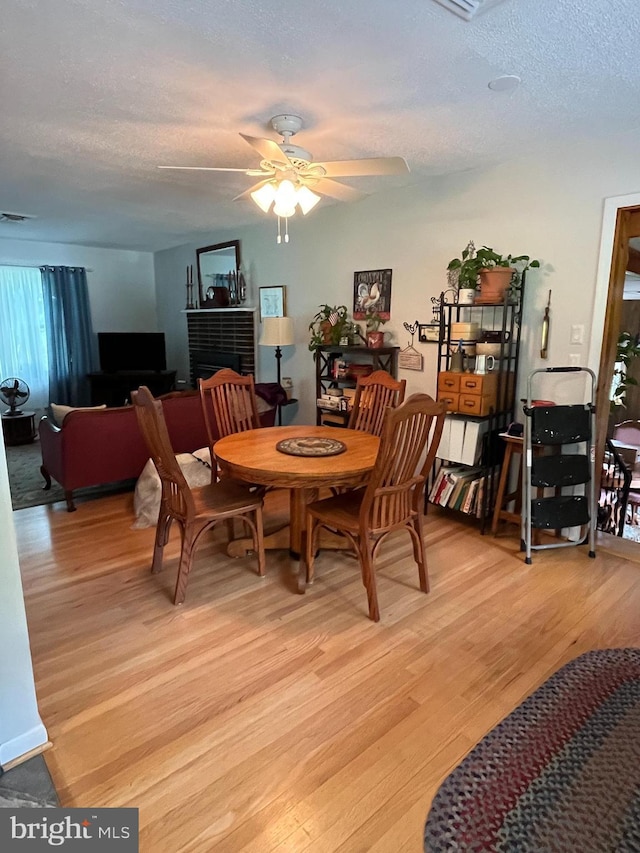 dining room featuring visible vents, ceiling fan, a textured ceiling, light wood-type flooring, and a brick fireplace