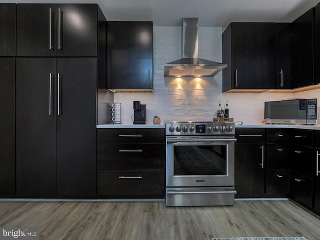 kitchen featuring light wood finished floors, wall chimney range hood, stainless steel electric range, and dark cabinetry