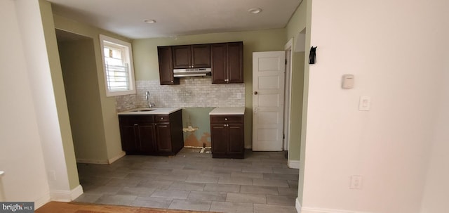 kitchen featuring dark brown cabinetry, under cabinet range hood, baseboards, light countertops, and backsplash