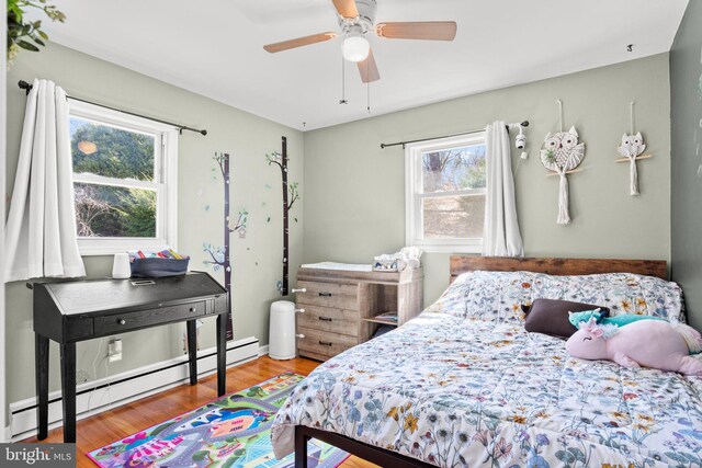 bedroom featuring a baseboard heating unit, multiple windows, wood finished floors, and a ceiling fan