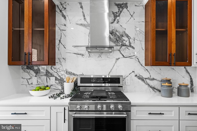 kitchen with wall chimney exhaust hood, glass insert cabinets, stainless steel gas range, light countertops, and backsplash