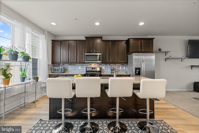 kitchen featuring light stone counters, a center island with sink, stainless steel appliances, tasteful backsplash, and dark brown cabinetry