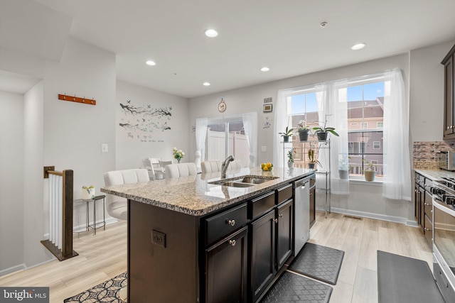 kitchen with light stone counters, a sink, appliances with stainless steel finishes, light wood finished floors, and a center island with sink