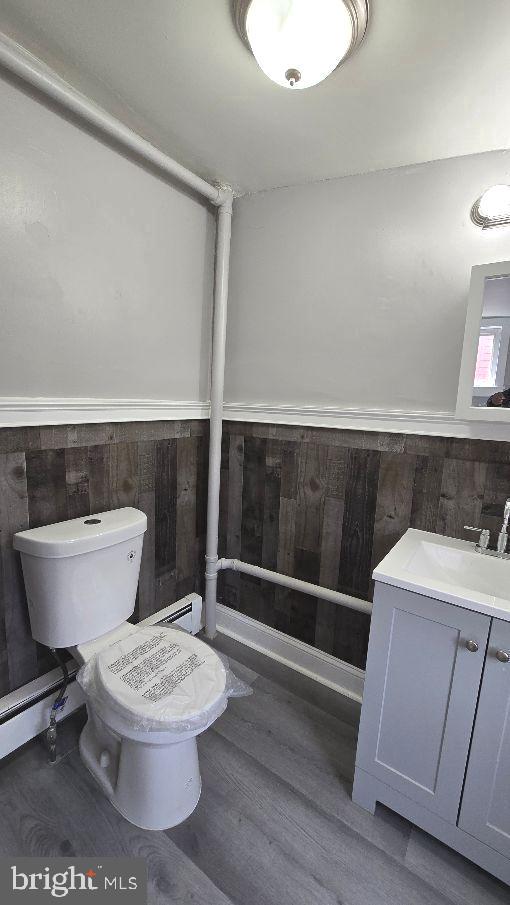 half bath featuring toilet, a wainscoted wall, wood finished floors, vanity, and a baseboard heating unit