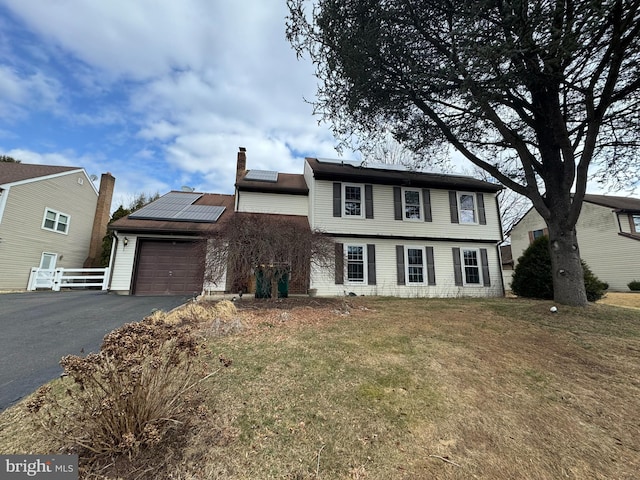 colonial-style house with aphalt driveway, an attached garage, solar panels, fence, and a chimney