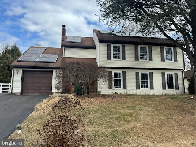 colonial-style house featuring a garage, a chimney, aphalt driveway, roof mounted solar panels, and a front yard