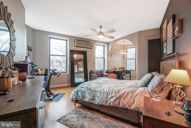 bedroom featuring arched walkways, light wood finished floors, a wall mounted AC, ceiling fan, and baseboards