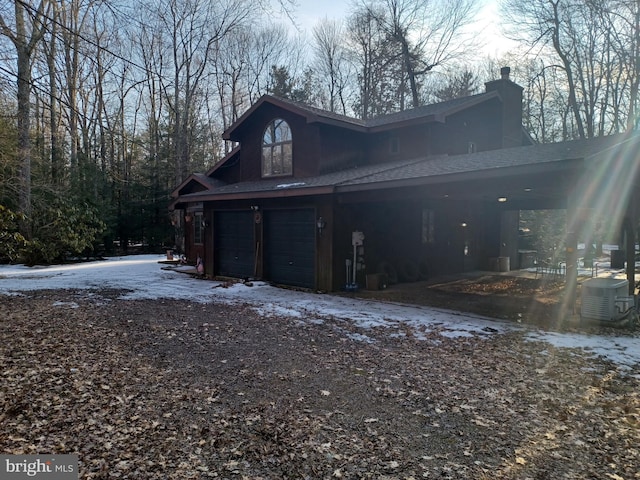 view of snow covered exterior with aphalt driveway, a chimney, an attached garage, and cooling unit