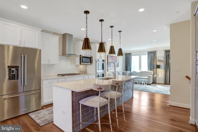 kitchen featuring a kitchen island with sink, a sink, wood finished floors, appliances with stainless steel finishes, and wall chimney exhaust hood