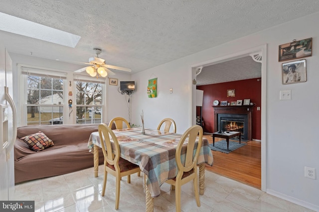 dining room with a textured ceiling, ceiling fan, a skylight, baseboards, and a lit fireplace