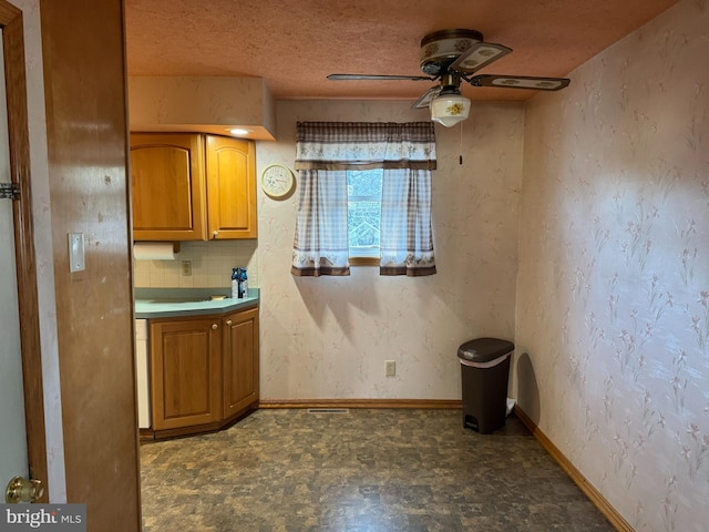 kitchen featuring a textured ceiling, a ceiling fan, baseboards, brown cabinets, and wallpapered walls