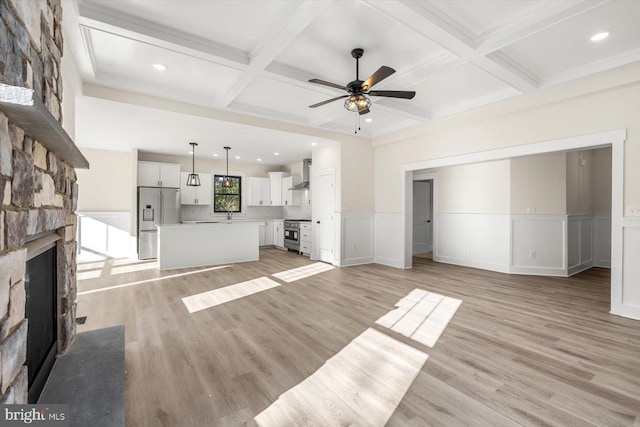 unfurnished living room featuring light wood finished floors, a stone fireplace, a wainscoted wall, and coffered ceiling