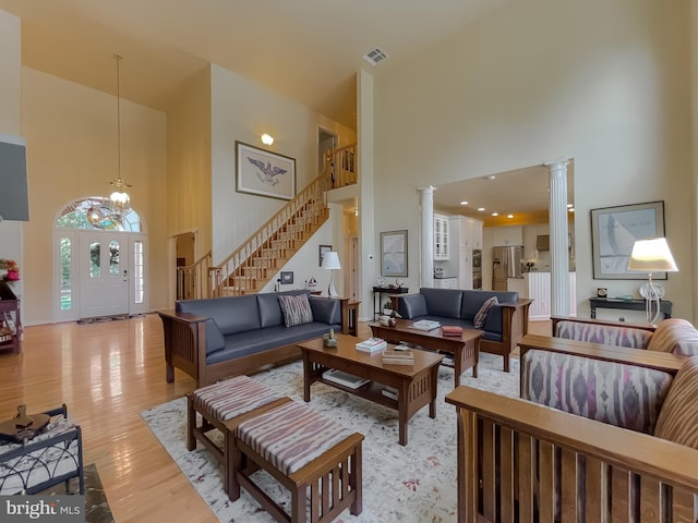 living room with a chandelier, stairs, light wood-style flooring, a towering ceiling, and ornate columns