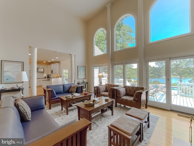 living room with light wood-style flooring, a towering ceiling, a water view, and ornate columns