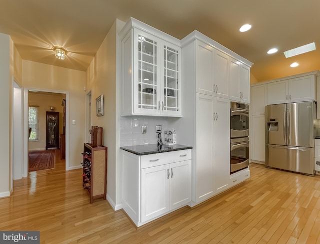 kitchen with light wood-style flooring, stainless steel appliances, white cabinets, glass insert cabinets, and backsplash