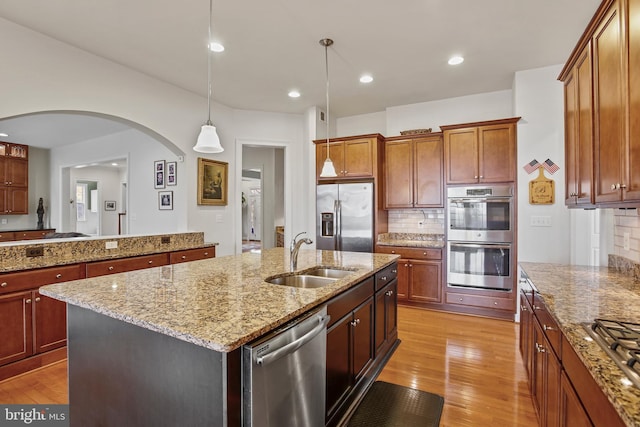 kitchen with an island with sink, a sink, stainless steel appliances, light wood-style floors, and light stone countertops