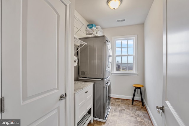 laundry area featuring baseboards, visible vents, stacked washing maching and dryer, cabinet space, and stone finish floor