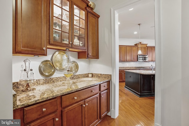 kitchen with light wood finished floors, stainless steel microwave, tasteful backsplash, glass insert cabinets, and light stone counters