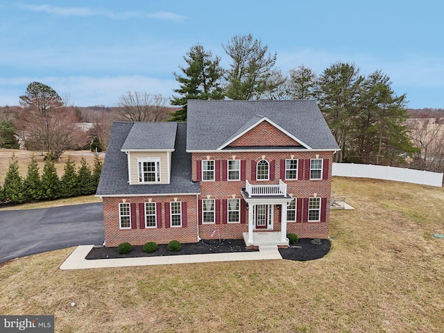 colonial inspired home with aphalt driveway, roof with shingles, fence, a front lawn, and brick siding