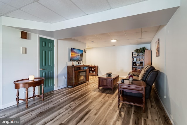 living area featuring wood finished floors, a glass covered fireplace, a paneled ceiling, and baseboards