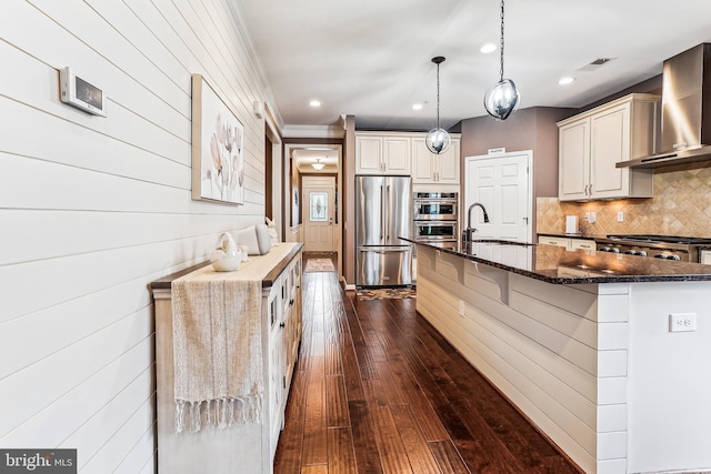 kitchen featuring backsplash, wall chimney range hood, dark stone countertops, appliances with stainless steel finishes, and a sink