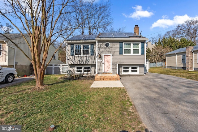 split foyer home with fence, a chimney, a front lawn, aphalt driveway, and roof mounted solar panels