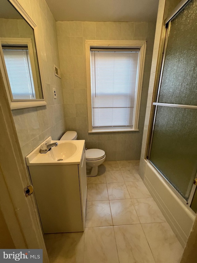 full bath featuring tile patterned flooring, toilet, bath / shower combo with glass door, vanity, and tile walls