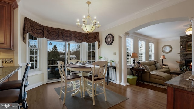 dining room with arched walkways, a stone fireplace, crown molding, dark wood-type flooring, and ornate columns