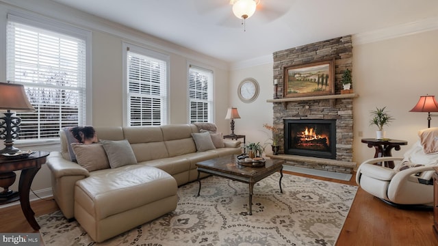 living room with a ceiling fan, crown molding, a stone fireplace, and wood finished floors