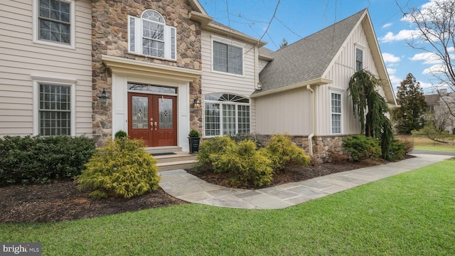 entrance to property with a shingled roof, stone siding, french doors, a lawn, and board and batten siding